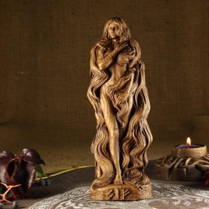May include: A wooden sculpture of a woman with long flowing hair. The sculpture is carved in a detailed style and has a natural wood finish. The woman is standing with her arms crossed in front of her. The sculpture is about 12 inches tall.
