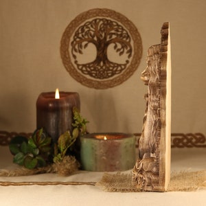 May include: A wooden wall decor featuring a carved image of a woman with long hair and flowing robes. The decor is in a natural wood color and has a detailed design. The decor is displayed in front of a brown background with candles and greenery.