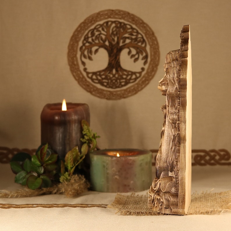 May include: A wooden wall decor featuring a carved image of a woman with long hair and flowing robes. The decor is in a natural wood color and has a detailed design. The decor is displayed in front of a brown background with candles and greenery.
