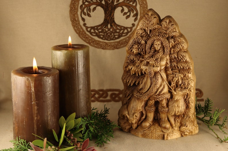 May include: A wooden carving of a woman standing in a forest, holding a staff. She is surrounded by trees and a wolf. The carving is on a wooden base with the word "KENI" inscribed on it.