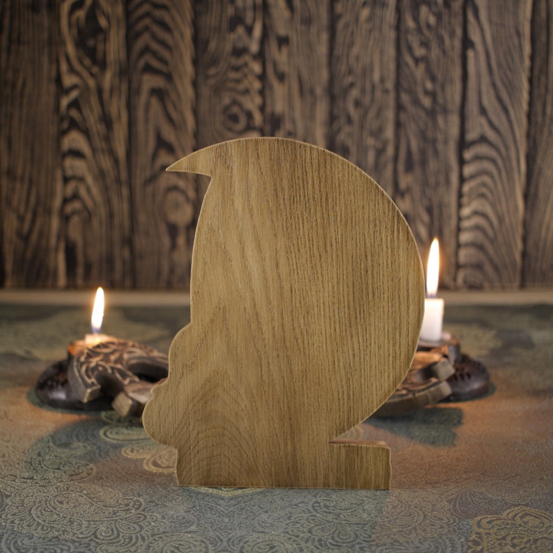 May include: A wooden cutout of a person's head with a rounded shape. The cutout is light brown and has a smooth surface. It is sitting on a patterned surface with two lit candles in the background.
