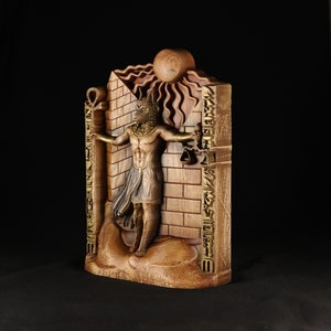 May include: A wooden sculpture of an Egyptian god with a jackal head, standing in front of a brick wall with hieroglyphs. The god is holding a staff in each hand and is surrounded by a sunburst.