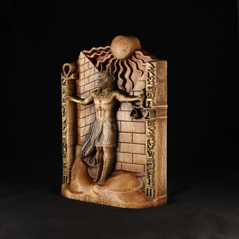 May include: A wooden sculpture of an Egyptian god with a jackal head, standing in front of a brick wall with hieroglyphs. The god is holding a staff in each hand and is surrounded by a sunburst.