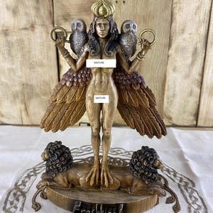 Könnte beinhalten: Holzstatue einer geflügelten weiblichen Figur mit Eulen auf den Schultern und Löwen zu ihren Füßen. Die Figur ist nackt und trägt eine Krone auf dem Kopf. Die Statue ist in Braun- und Goldtönen bemalt.