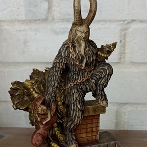 May include: A detailed figurine of Krampus, a horned figure with a fur coat and chains, perched atop a brick structure. The figure holds a small Christmas tree and a person. The statue is painted in shades of brown, tan, and gold.
