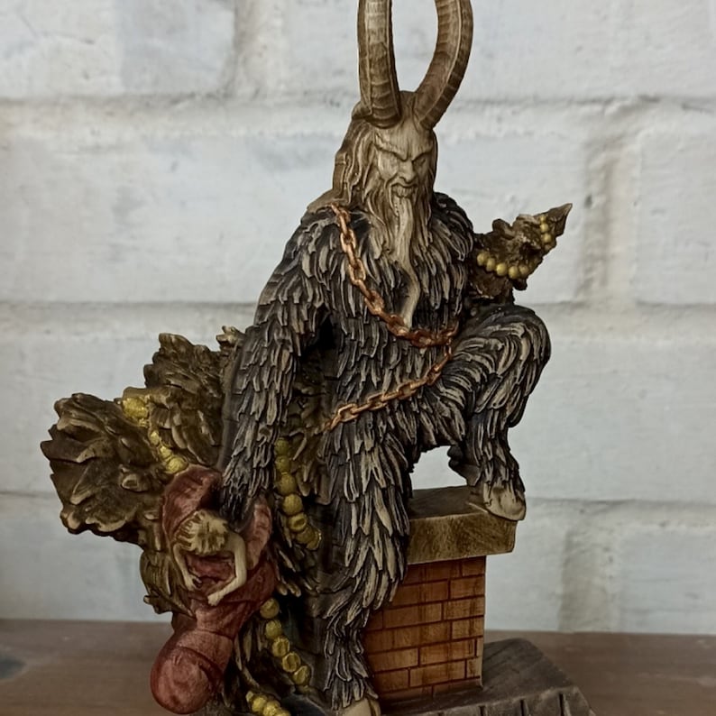 May include: A detailed figurine of Krampus, a horned figure with a fur coat and chains, perched atop a brick structure. The figure holds a small Christmas tree and a person. The statue is painted in shades of brown, tan, and gold.