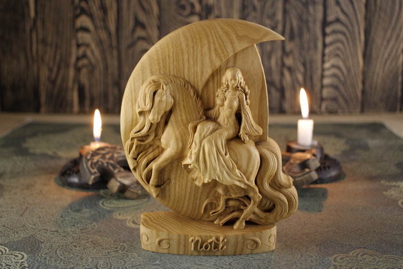 May include: A wooden carving of a woman riding a horse, set against a crescent moon background. The carving is light brown and has intricate details. The woman is wearing a long flowing dress and has long flowing hair. The horse has a flowing mane and tail. The carving is signed "Nott" at the base.
