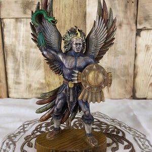 Op de afbeelding: Een gedetailleerd beeldje van de godheid Huitzilopochtli, met een gespierd figuur met grote gevederde vleugels, een groene slang in de rechterhand en een rond schild in de linkerhand. Het figuur draagt een hoofdtooi en staat op een houten voet.