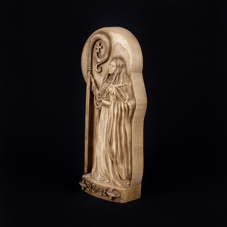 May include: A wooden carving of Saint Brigid, a female saint, holding a staff with a cross. The carving is on a wooden plaque with the words "St Brigid" inscribed at the bottom.