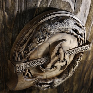 May include: A wooden wall plaque with a carved image of two wolves flanking a triquetra symbol. The wolves are depicted in a stylized, almost mythical way, with their paws and tails intertwined. The triquetra is a Celtic knot symbol that represents the trinity, and it is often used in pagan and Wiccan traditions.