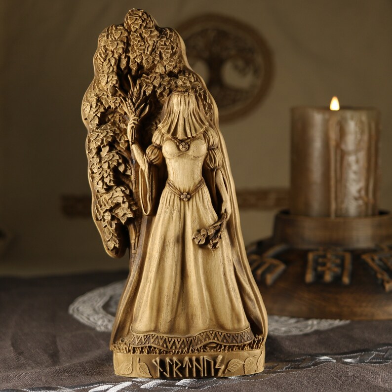 Puede incluir: Una estatua de madera de la diosa n&oacute;rdica Freyja, de pie frente a un &aacute;rbol. Lleva un vestido largo y fluido y un velo. La estatua est&aacute; inscrita con la palabra "Freyja".