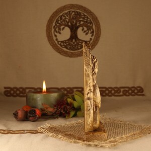 May include: A wooden carving of a Norse god, possibly Thor, with a hammer in his hand. The carving is on a wooden base and is painted in a gold color. The carving is in front of a candle and a tree of life symbol.
