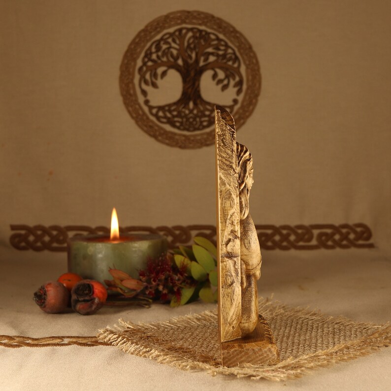 May include: A wooden carving of a Norse god, possibly Thor, with a hammer in his hand. The carving is on a wooden base and is painted in a gold color. The carving is in front of a candle and a tree of life symbol.
