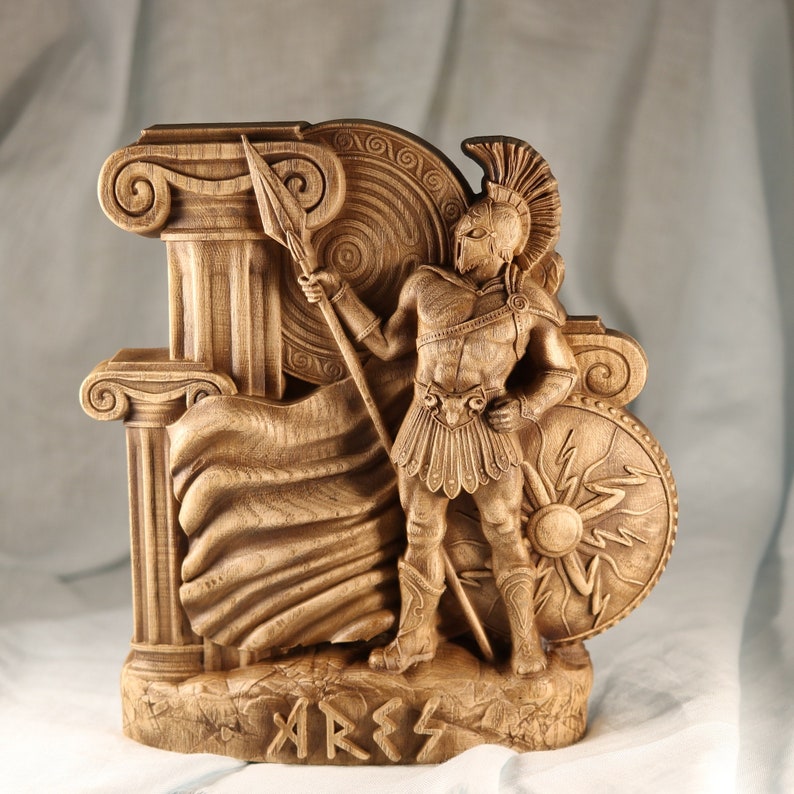 A wooden sculpture of the Greek god Ares, depicted as a warrior holding a spear and shield. The sculpture is detailed with intricate carvings and features a flowing cape and a base inscribed with the word "Ares".