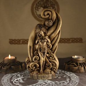 Op de afbeelding: Een houten sculptuur van een godinfiguur die in een wervelend wolkachtig ontwerp staat. De godin houdt een hart in haar handen en heeft een kroon op haar hoofd. Het beeld is bruin en heeft een natuurlijke houten afwerking.