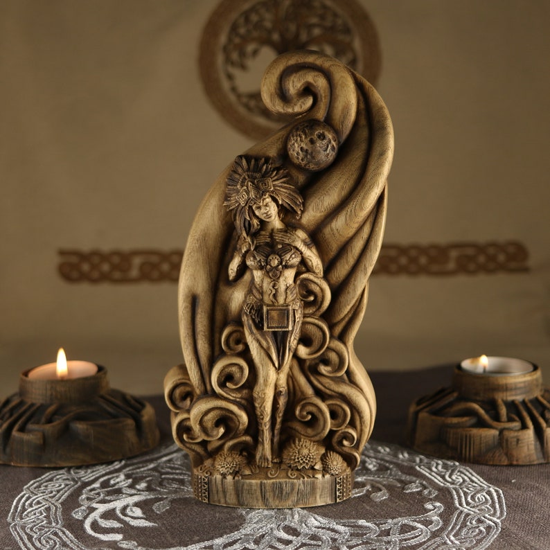 Op de afbeelding: Een houten sculptuur van een godinfiguur die in een wervelend wolkachtig ontwerp staat. De godin houdt een hart in haar handen en heeft een kroon op haar hoofd. Het beeld is bruin en heeft een natuurlijke houten afwerking.