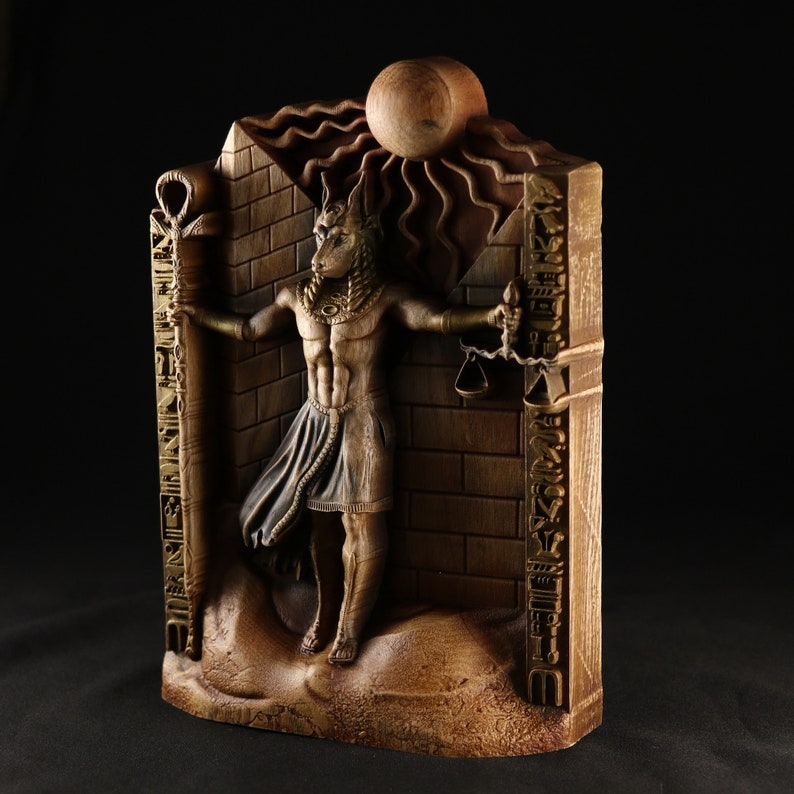 May include: A wooden sculpture of an Anubis figure holding a scale in one hand and a staff in the other. The sculpture is set against a brick wall with hieroglyphics carved into the surface.
