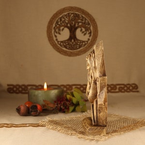 May include: A wooden carving of a woman with long hair, standing in a carved frame. The carving is painted in shades of brown and beige. The carving is on a burlap surface.