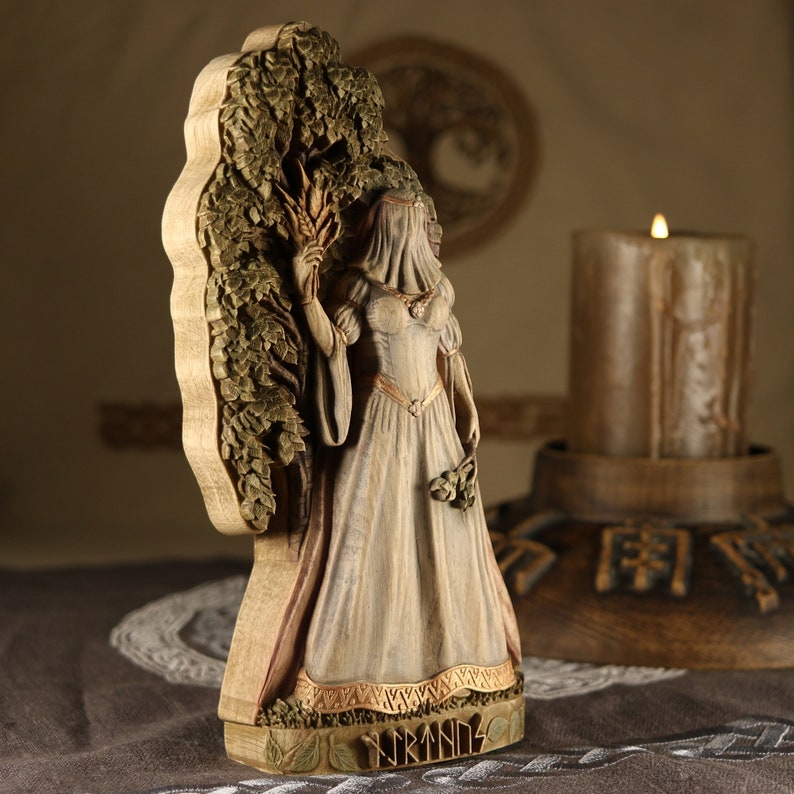 Puede incluir: Una escultura de madera de una mujer de pie frente a un &aacute;rbol. Lleva un vestido largo blanco y tiene un velo sobre la cabeza. La escultura est&aacute; pintada en tonos marrones y blancos. La escultura est&aacute; etiquetada como "Yrmins".