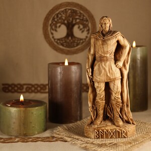 May include: A wooden statue of a Viking warrior with a beard and a cloak, standing on a wooden base with runes. The statue is surrounded by three candles, two brown and one green.