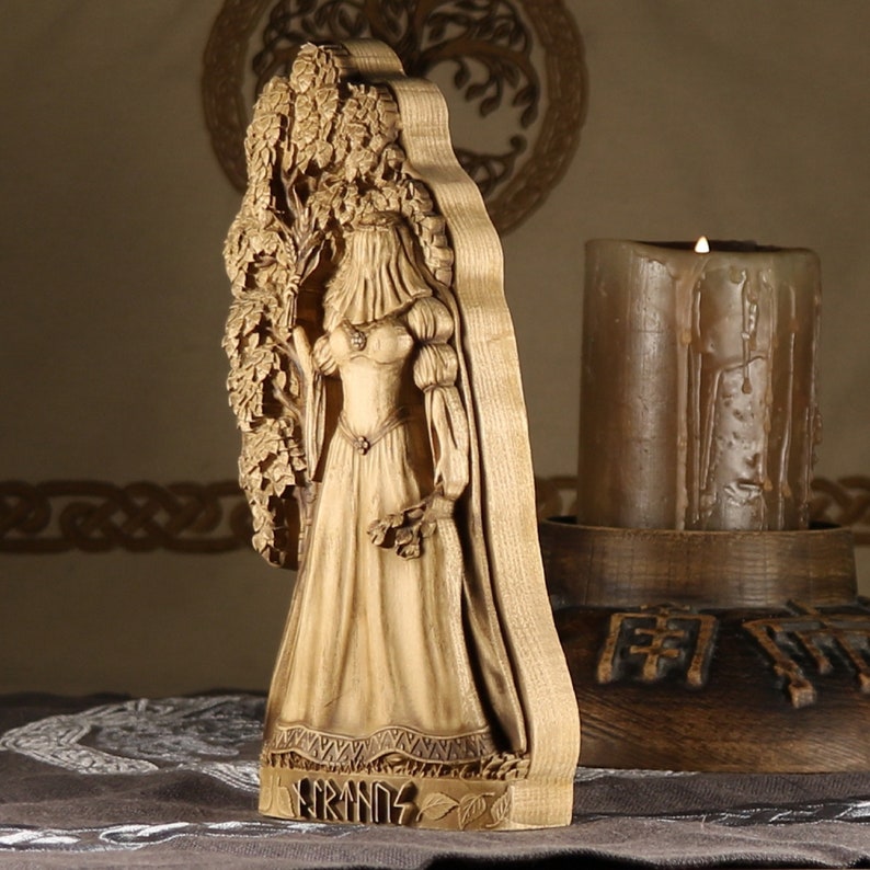 Puede incluir: Una talla de madera de la diosa n&oacute;rdica Freyja, de pie frente a un &aacute;rbol. Lleva un vestido largo y un velo. La talla est&aacute; sobre una base de madera con la palabra "Freyja" inscrita en ella.