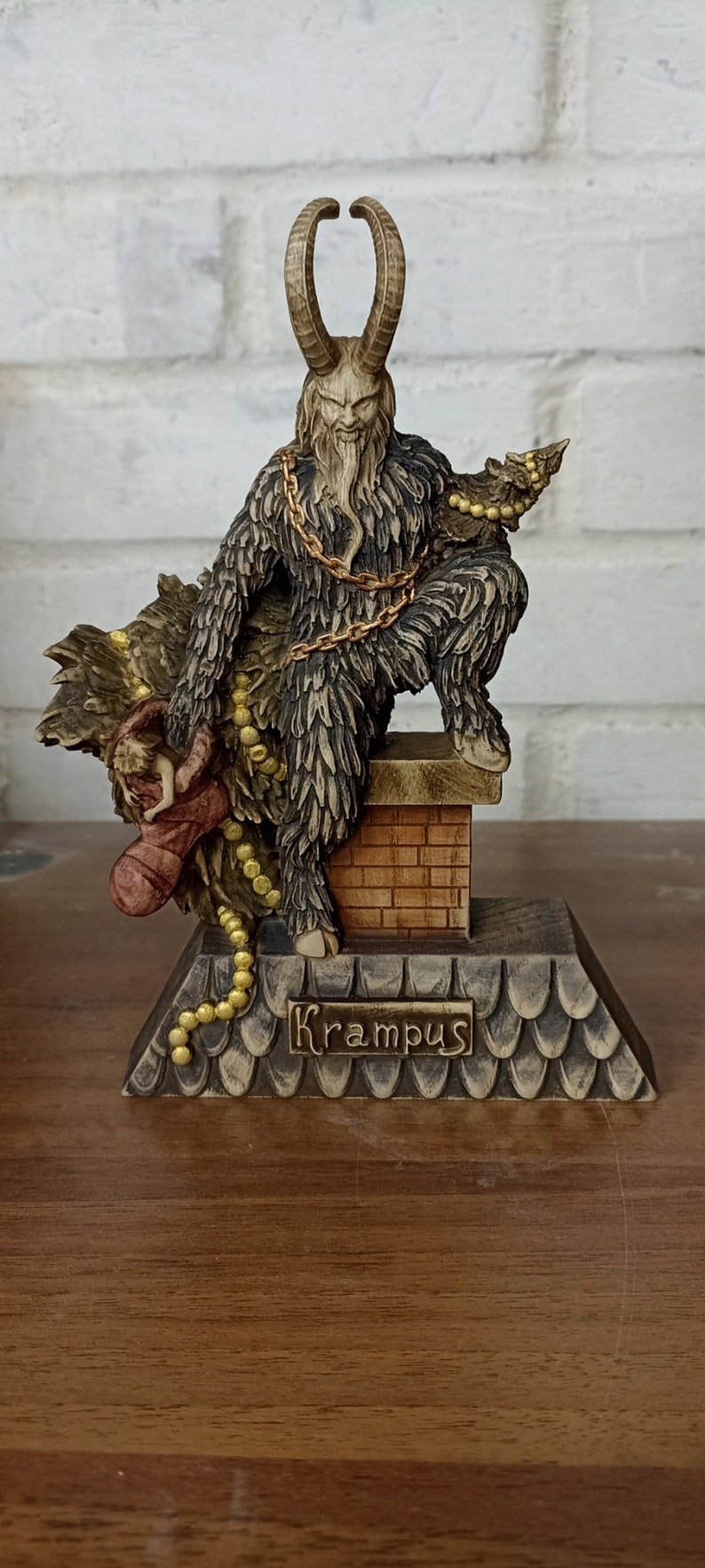 May include: A Krampus figurine with a horned head, shaggy fur, and a gold chain. The figure is perched on a chimney, holding a sack and a string of beads. The base is inscribed with the name "Krampus".