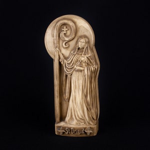 May include: A wooden carving of Saint Brigid, a female saint, holding a crosier. The carving is painted in shades of brown and has a halo around the saint's head. The carving is labeled "St Brigid" at the bottom.