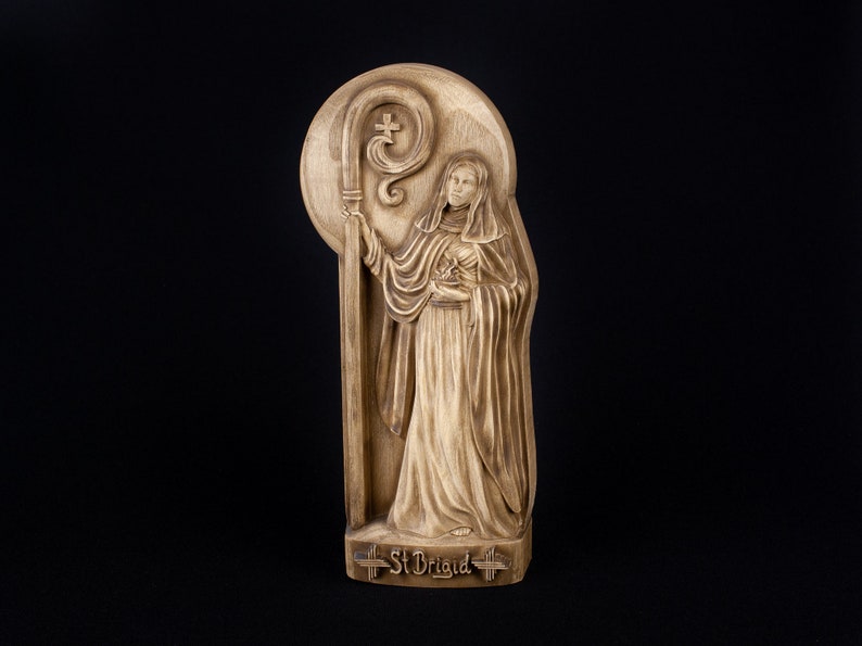 May include: A wooden carving of Saint Brigid, a female saint, holding a crosier. The carving is painted in shades of brown and has a halo around the saint's head. The carving is labeled "St Brigid" at the bottom.