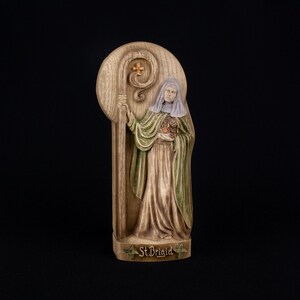 May include: A wooden plaque with a relief sculpture of a woman holding a staff and a heart. The woman is wearing a green robe and a white head covering. The plaque is inscribed with the words "St. Brigid".