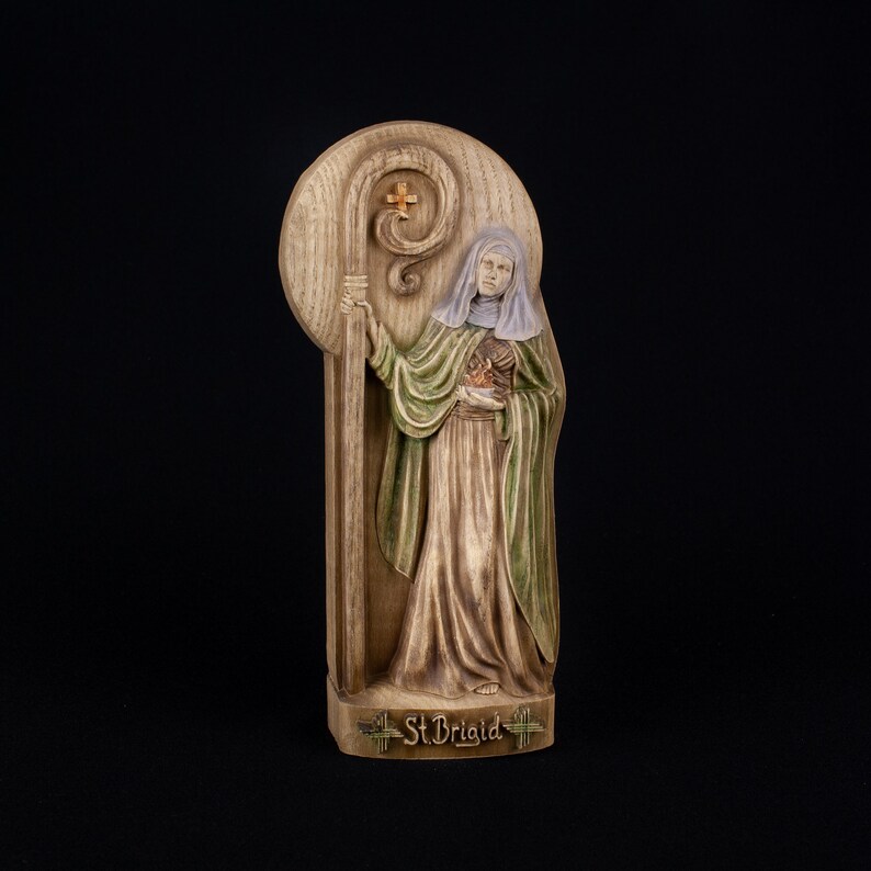 May include: A wooden plaque with a relief sculpture of a woman holding a staff and a heart. The woman is wearing a green robe and a white head covering. The plaque is inscribed with the words "St. Brigid".