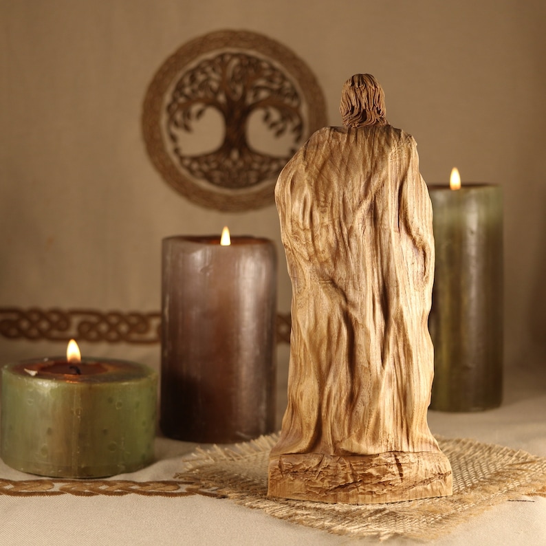 May include: A wooden sculpture of a person wearing a long robe, standing in front of three lit candles. The candles are in different colors, green, brown, and gray. The sculpture is on a burlap surface.