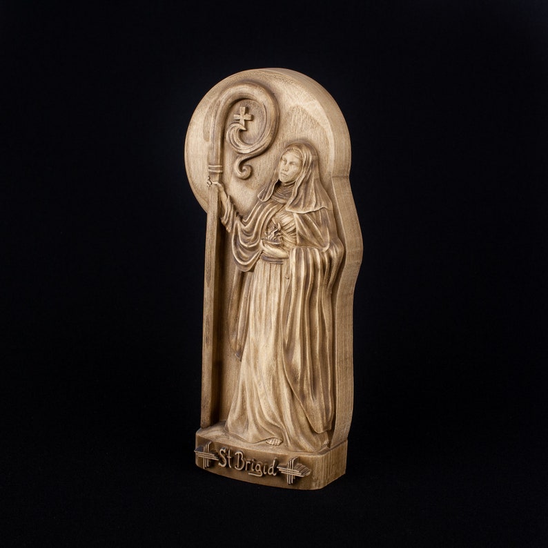 May include: A wooden carving of Saint Brigid, a female saint, holding a crosier. The carving is on a wooden plaque with the words "St Brigid" inscribed at the bottom.