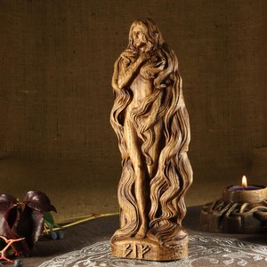 May include: A wooden sculpture of a woman with long flowing hair. The sculpture is carved in a detailed style and has a natural wood finish. The sculpture is standing on a base with the rune "Bjarkan" carved into it.