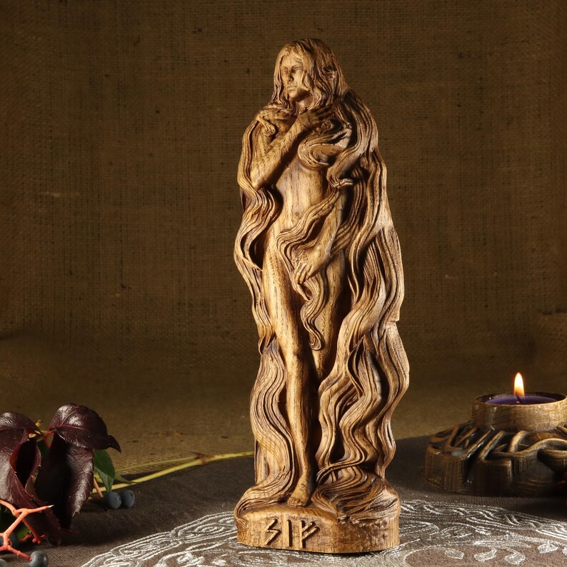 May include: A wooden sculpture of a woman with long flowing hair. The sculpture is carved in a detailed style and has a natural wood finish. The sculpture is standing on a base with the rune "Bjarkan" carved into it.