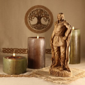 May include: A wooden carving of a Viking warrior standing on a base with runes. The warrior is wearing a cloak and holding a sword. The carving is surrounded by three candles.