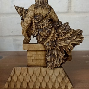 May include: A detailed wooden sculpture of a horned figure with a fur-like cloak and a decorative skirt. The figure is perched on a small, tiled roof structure. The sculpture is a warm, natural wood color.