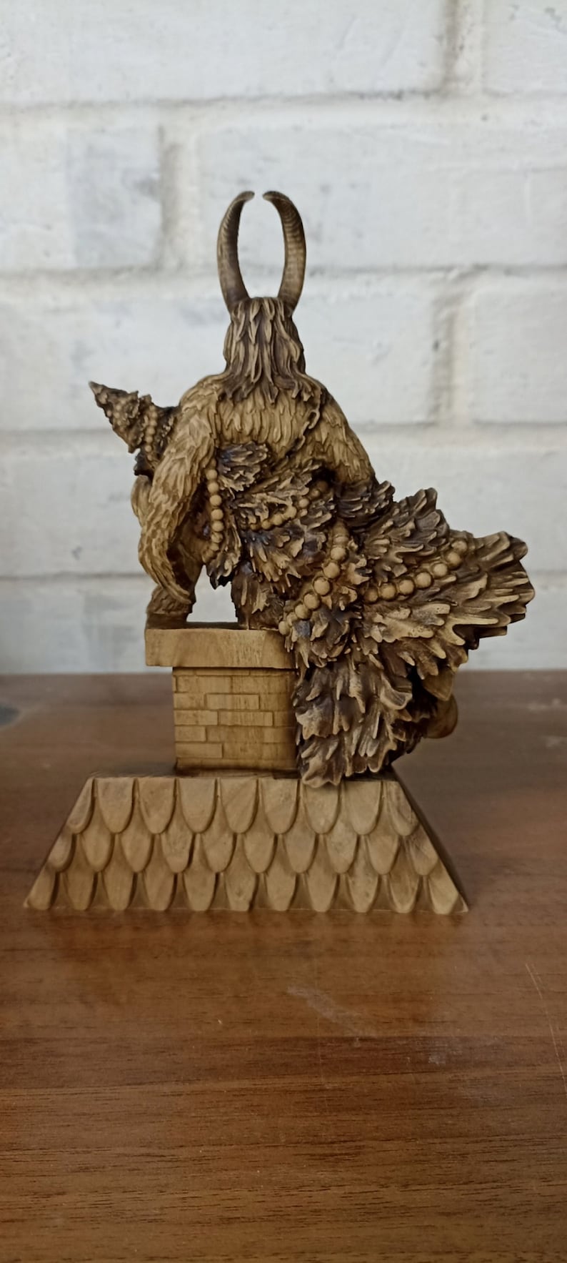May include: A detailed wooden sculpture of a horned figure with a fur-like cloak and a decorative skirt. The figure is perched on a small, tiled roof structure. The sculpture is a warm, natural wood color.