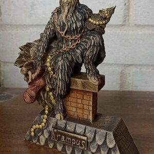 May include: A detailed figurine of Krampus, a horned figure from Alpine folklore, perched atop a chimney. The statue features a brown and black color scheme, with gold accents and the name "Krampus" inscribed on the base.