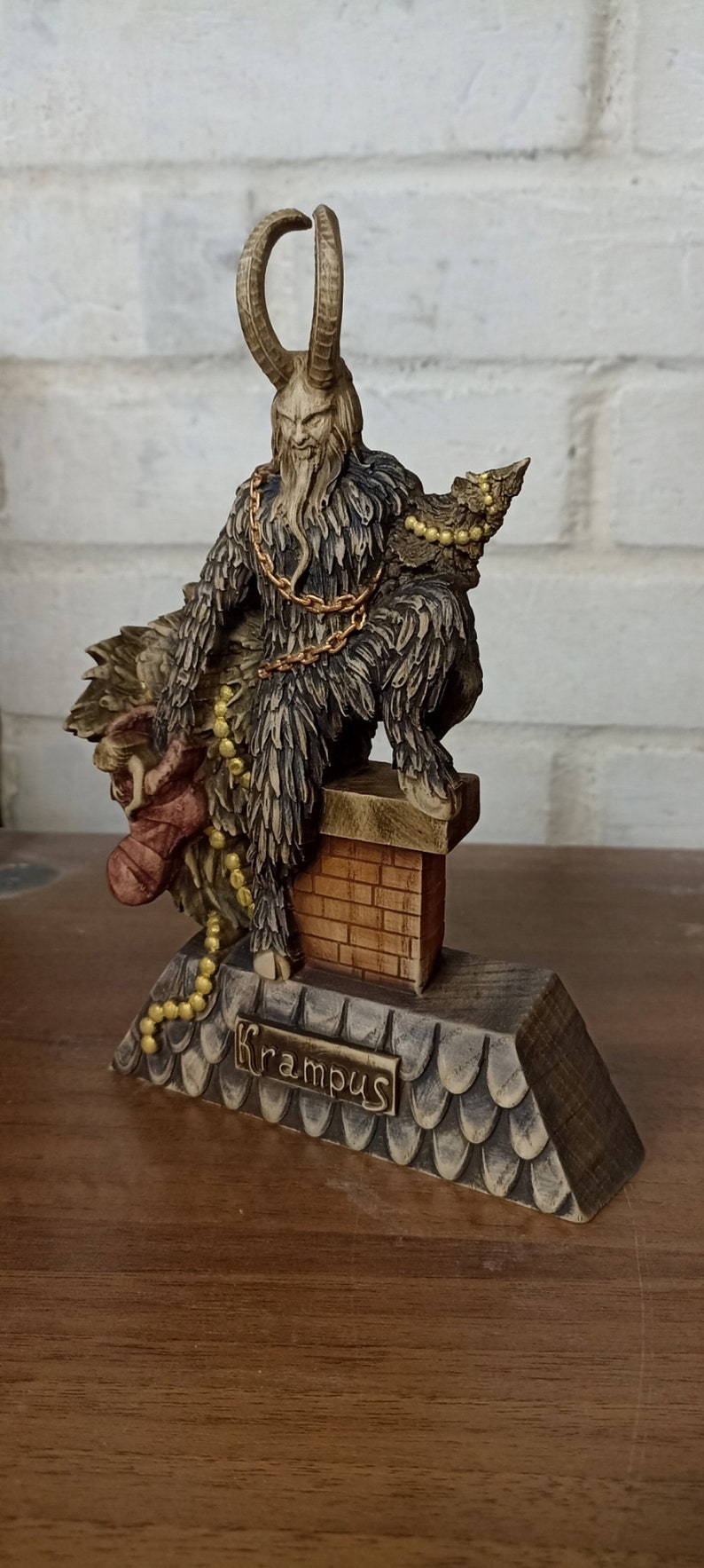 May include: A detailed figurine of Krampus, a horned figure from Alpine folklore, perched atop a chimney. The statue features a brown and black color scheme, with gold accents and the name "Krampus" inscribed on the base.