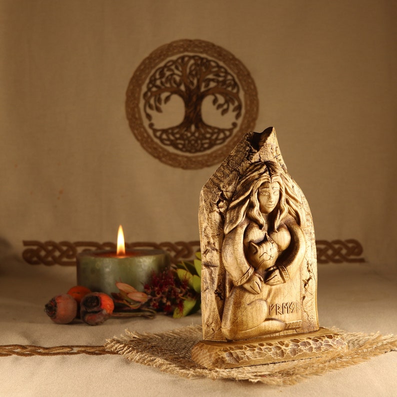 May include: Wooden carving of a woman holding a cat, with the word "FRIGG" carved below. The carving is on a wooden base and is in front of a brown background with a Celtic knot design.