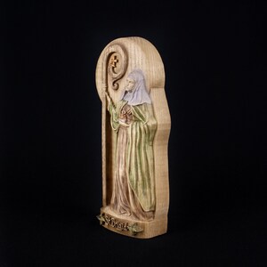 May include: A wooden carving of Saint Brigid, a female saint, holding a staff with a cross. The carving is painted in shades of brown, green, and white. The carving is labeled "St. Brigid" at the bottom.