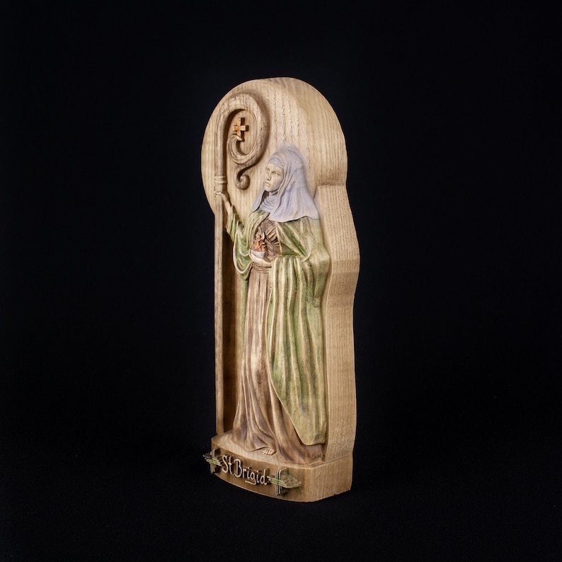 May include: A wooden carving of Saint Brigid, a female saint, holding a staff with a cross. The carving is painted in shades of brown, green, and white. The carving is labeled "St. Brigid" at the bottom.