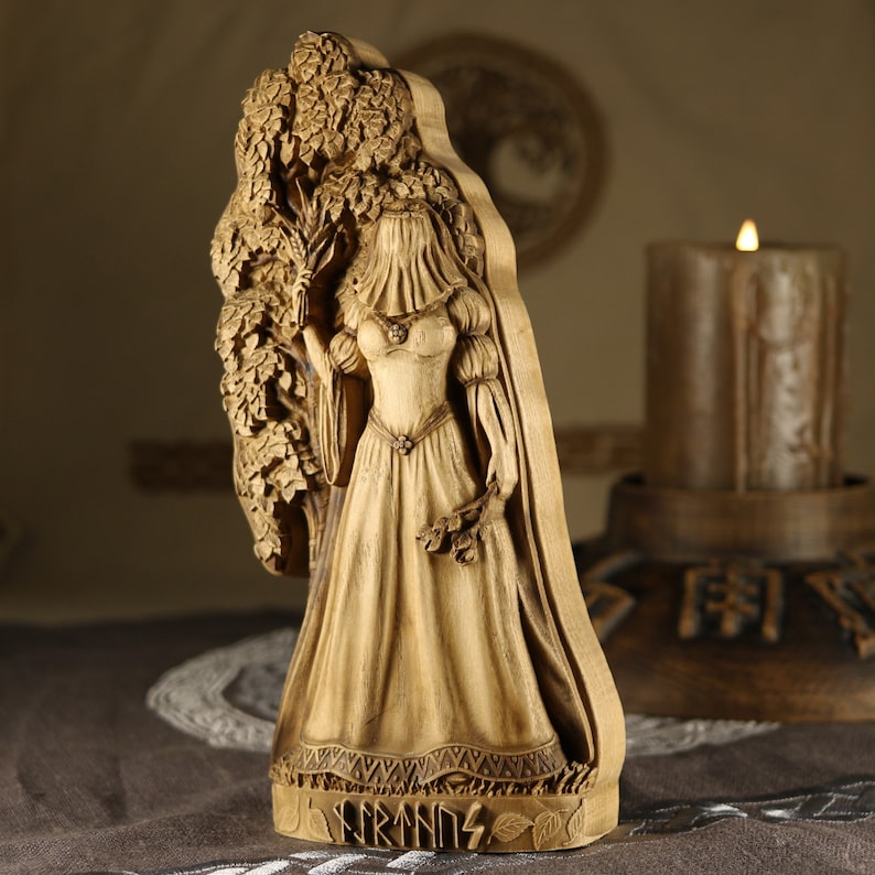 Puede incluir: Una talla de madera de una mujer con un vestido largo de pie junto a un &aacute;rbol. La talla est&aacute; pintada en tonos marrones y tiene la palabra "FRIGG" tallada en la parte inferior.
