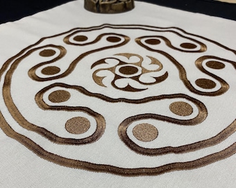 以下が含まれることがあります： 白い布に茶色の刺繍が施された円形のデザイン。デザインの中心には、4つのポイントと中央に円を持つ渦巻きがあります。渦巻きは、同心円とループのシリーズで囲まれています。