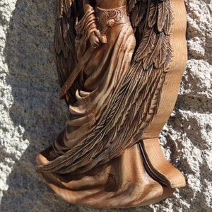 May include: A wooden carving of an angel with detailed wings and a flowing robe. The angel is holding a sword in its right hand.