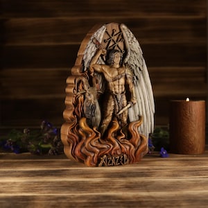 Op de afbeelding: Een houten sculptuur van een engel met vleugels, zwaard en dolk, boven vlammen. Een geit staat aan de voeten van de engel. De sculptuur heeft een donkerbruine afwerking en staat tegen een houten achtergrond.
