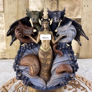 Op de afbeelding: Een houten beeldje van Tiamat, een vijfkoppige draakgodin uit de Babylonische mythologie. De godin is afgebeeld met een menselijke torso en vier drakenkoppen, elk in een andere kleur. Het beeldje is geschilderd in tinten bruin, goud en zwart. De basis van het beeldje is gegraveerd met het woord "Tiamat".