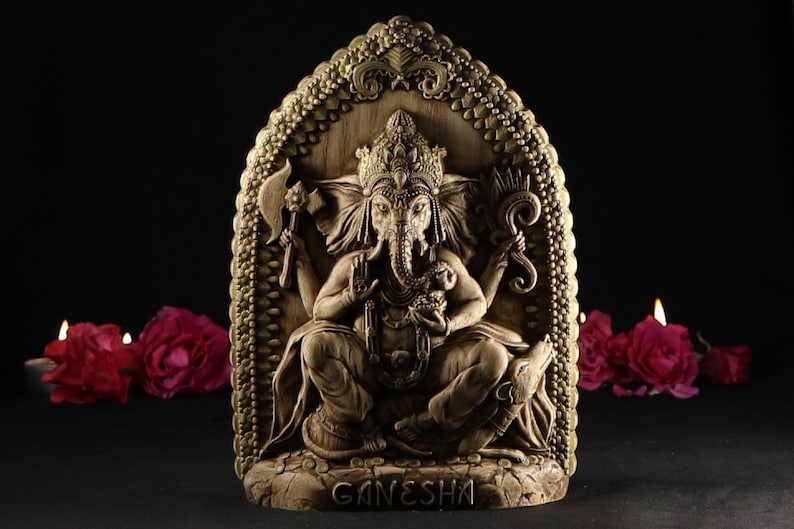 May include: A detailed, carved wooden sculpture of the Hindu deity Ganesha. The sculpture is painted in gold and brown tones and features intricate details. The sculpture is set against a black background with three lit candles and red roses in the foreground.