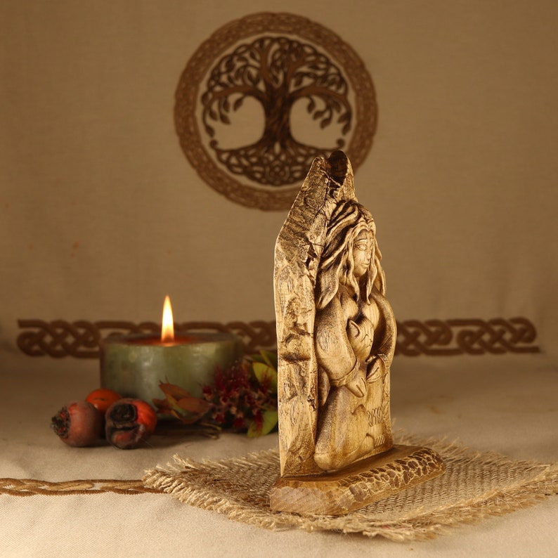 May include: A wooden carving of a woman holding a wolf cub. The carving is on a wooden base and is in front of a candle and a woven tapestry.