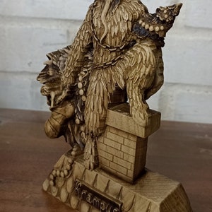 May include: A detailed, tan-colored Krampus figurine. The figure has large horns, a long beard, and is chained. The base is inscribed with the word "Krampus". The figurine is on a wooden surface.
